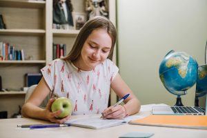 aluna segurando uma maçã e escrevendo no caderno com um globo atrás